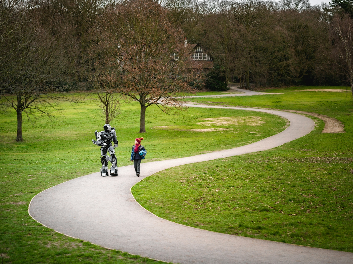 Photograph of a large robot walking and talking with a young boy in school uniform who's holding a football, down a winding path in the park.