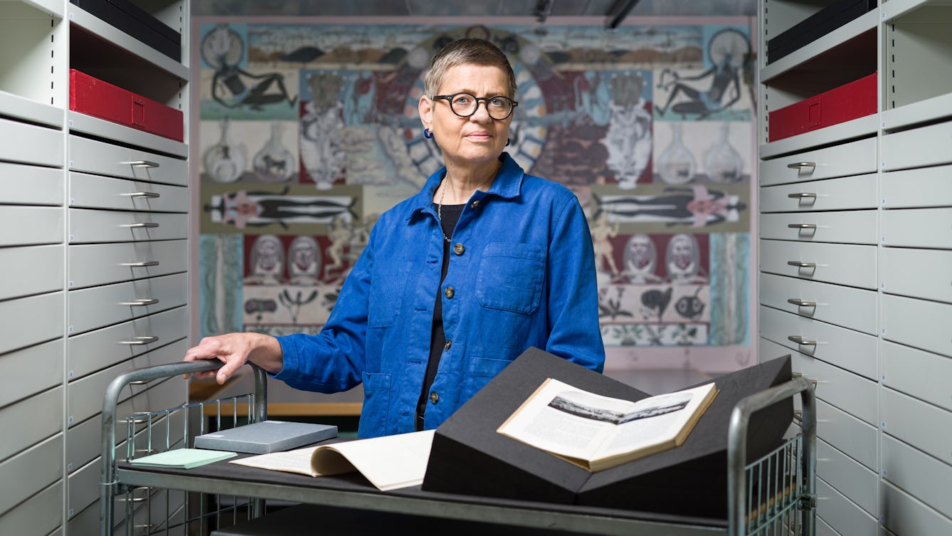 Photograph of a woman wearing a blue shirt standing behind a trolley in an archive store. One the trolley are open books and archive boxes. On either side of her are a series of thin drawers. In the background on the wall is a painted mural depicting various figures.