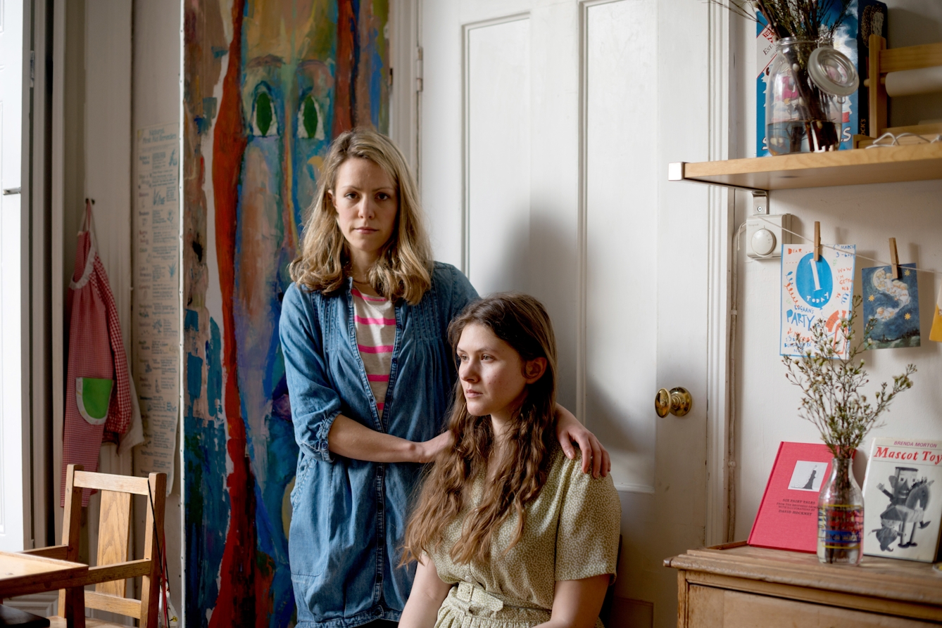 Photograph of a young woman and a mother figure in a living room. The young woman is seated in a long dress and and the mother figure stands behind her, hands on her shoulders. The young woman is looking off camera, the mother figure is looking to camera.