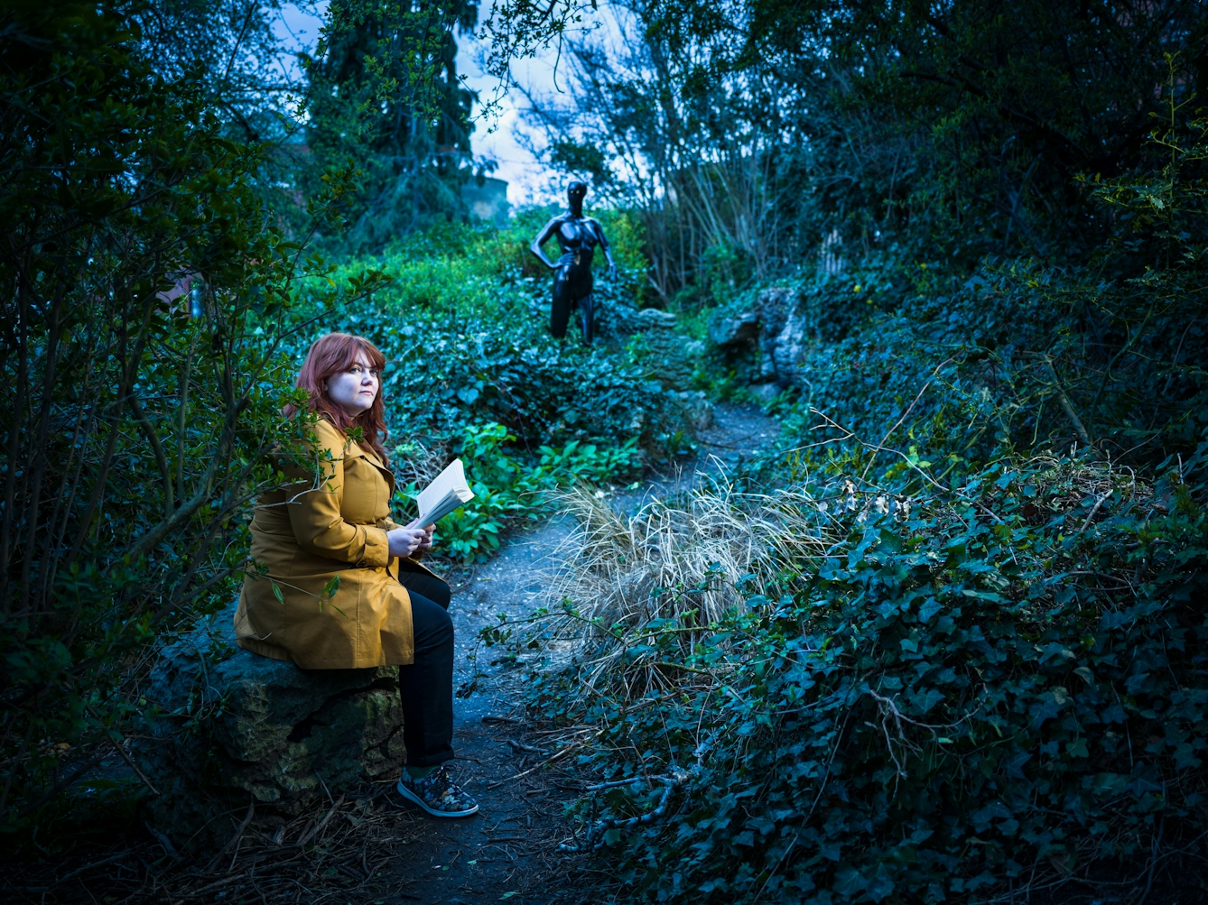 Photograph of a young woman sitting on a rock in a wooded area, looking up from reading her book. Behind her in the background a human form appears from the undergrowth.