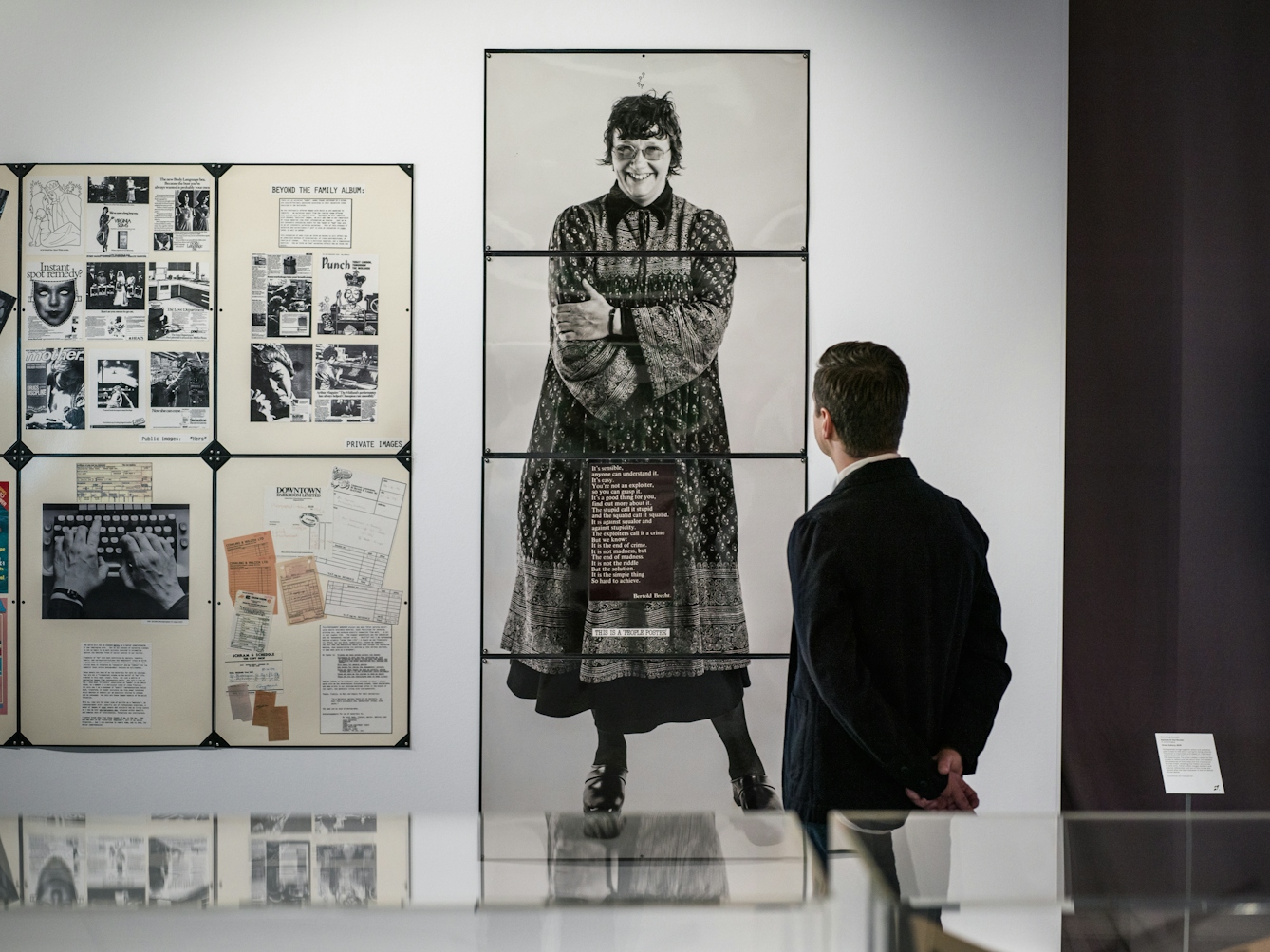 Photograph of a man in a dark jacket looking at a life-size picture of Jo Spence in the Misbehaving Bodies Exhibition at the Wellcome Collection. The man has his back to us whilst the picture of Jo Spence is gazing straight at the viewer of the picture. To the left of the photograph are various tear sheets from the work fo Jo Spence that make up 'Beyond the Family Album'.