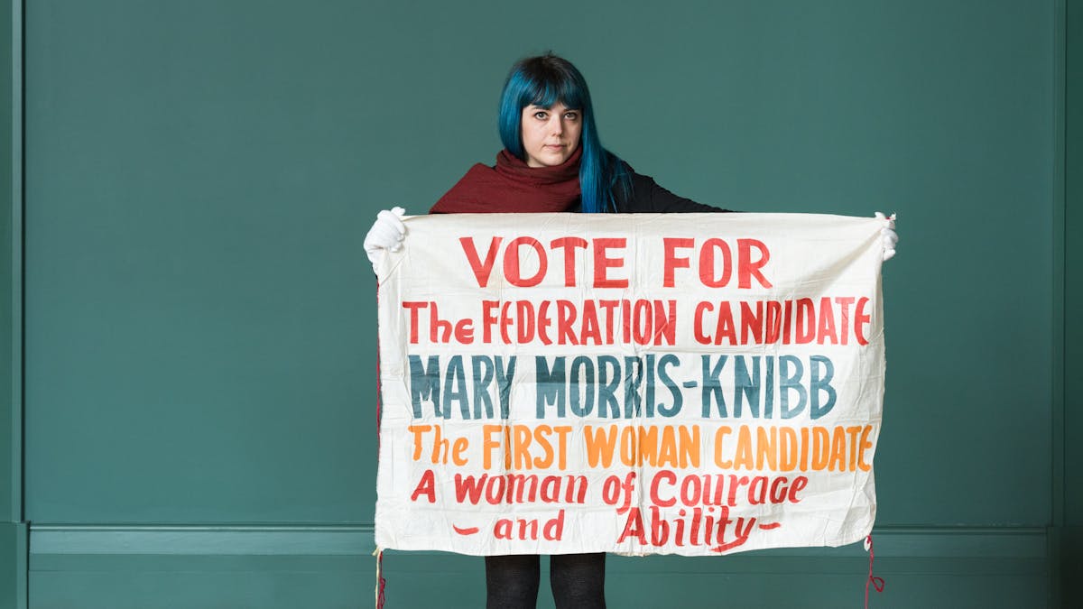 Photograph of a woman holding a 1930s cloth banner with the hand written words 'Vote for the Federation Candidate Mary Morris-Knibb the first woman candidate. A woman of courage and ability'.