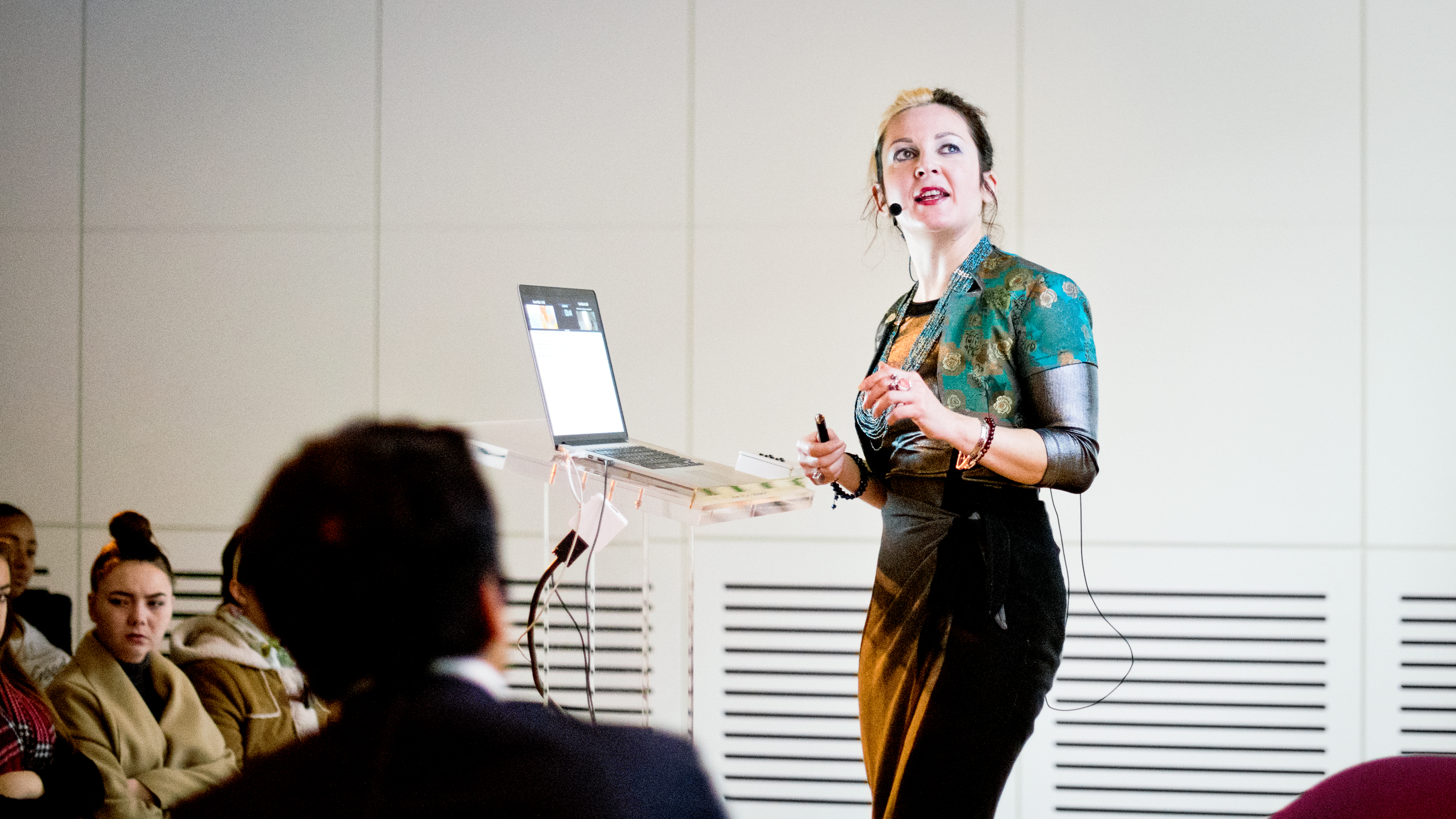 Photograph of a women with a head microphone standing at a lecturn with a laptop talks to an audience. The back of another panelist's head is out-of-focus in the foreground.