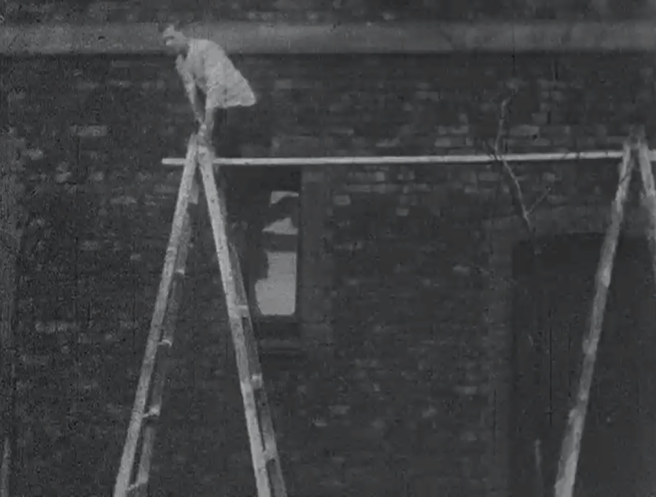 Black and white still showing a man with his hand bandaged up tightly, perched at the top of a ladder about to climb onto a plank balanced between his ladder and another.