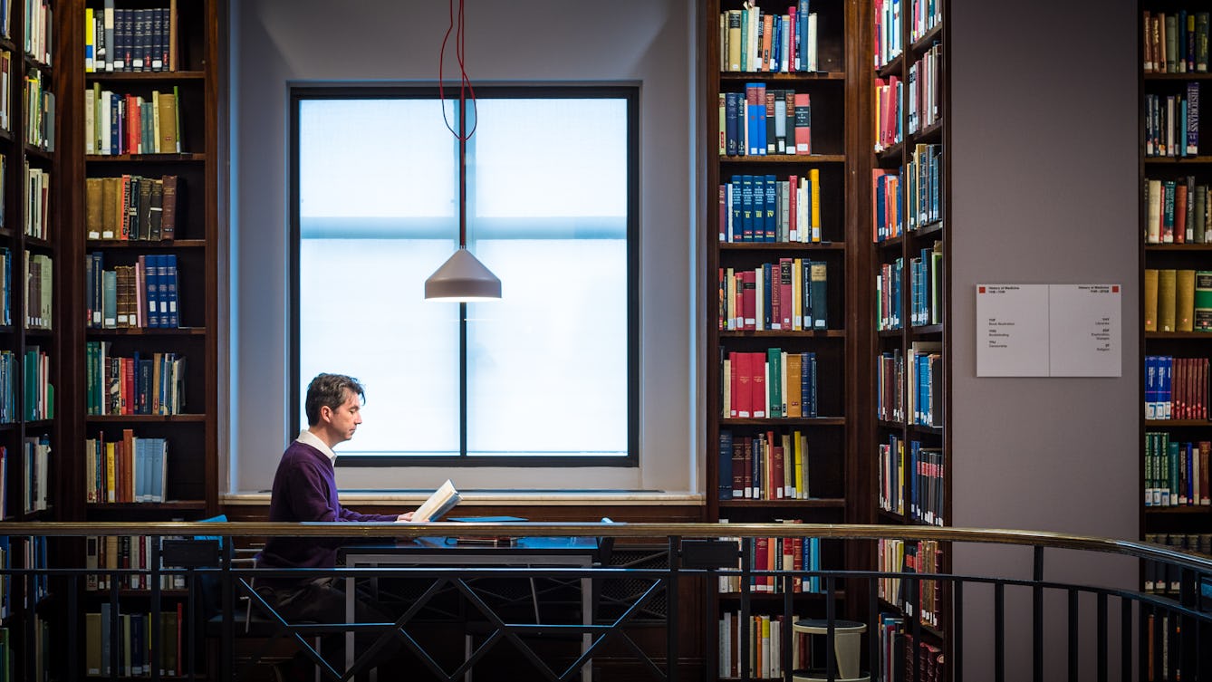 Photograph showing a man sat reading a book at a table in front of a window, surrounded by floor to ceiling  library shelves containing books.