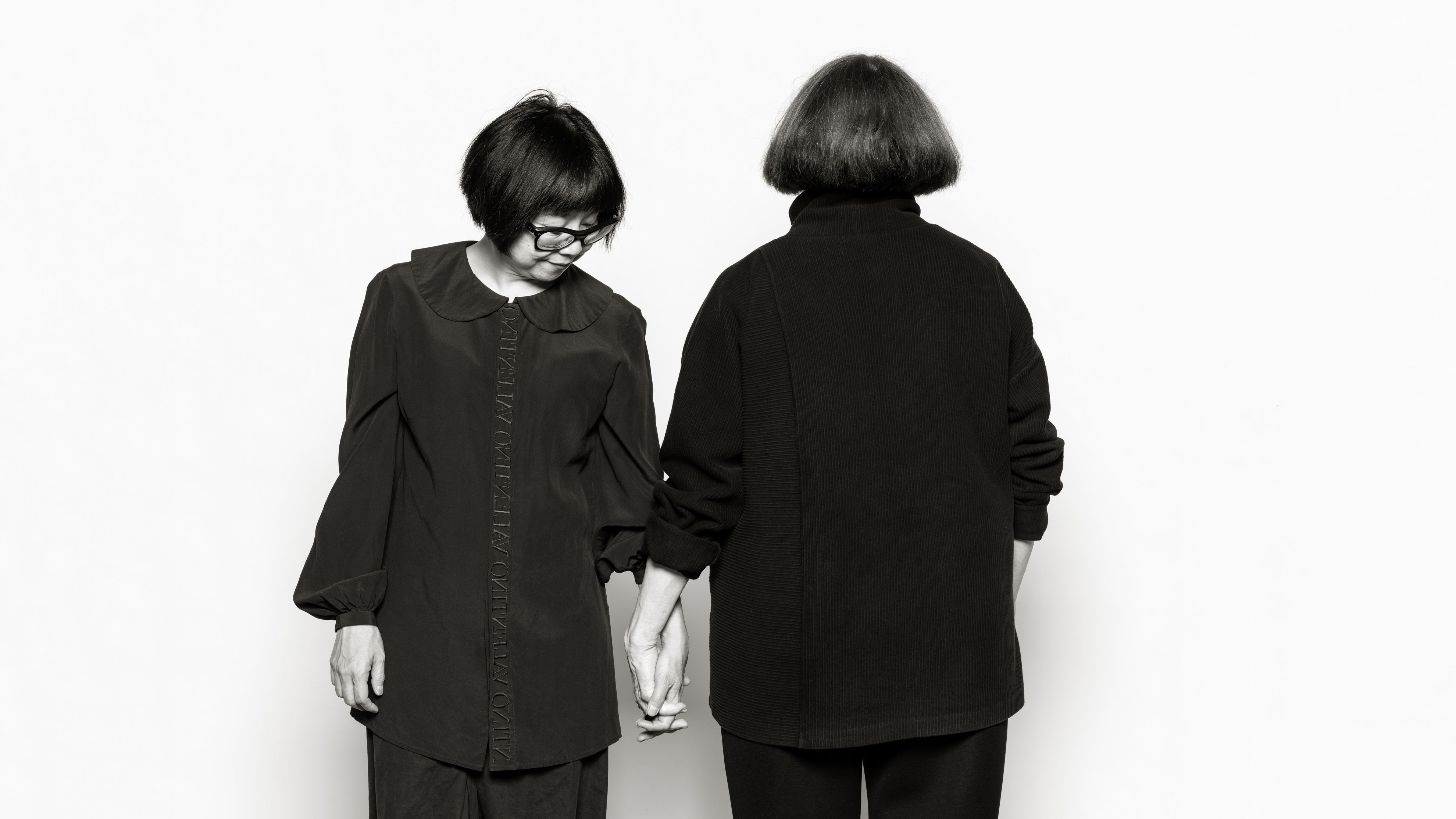 Photographic portrait of two women holding hands. One is facing away from the camera , the other is looking at their clasped hands.