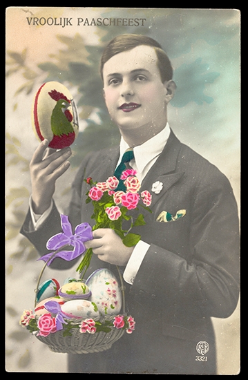 A young man in a smart suit holds an Easter egg with an image of a cockeral on it in one hand and a basket of decorated Easter eggs and a bunch of brightly coloured flowers in the other hand.