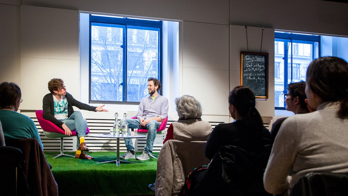 Photograph showing two young men sat in chairs on a small raised stage which is covered in artificial grass. One man is asking he other a question, with his hand and arm outstretched. The other man is smiling. In the foreground is the backs of the heads of the audience.