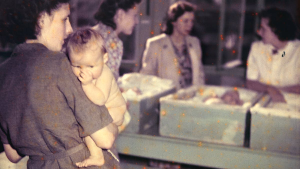 Photograph from the mid-20th Century Peckham Pioneer Centre, showing a women holding a baby in the foreground, and behind three women who appear to be conversing beside three cribs containing babies.