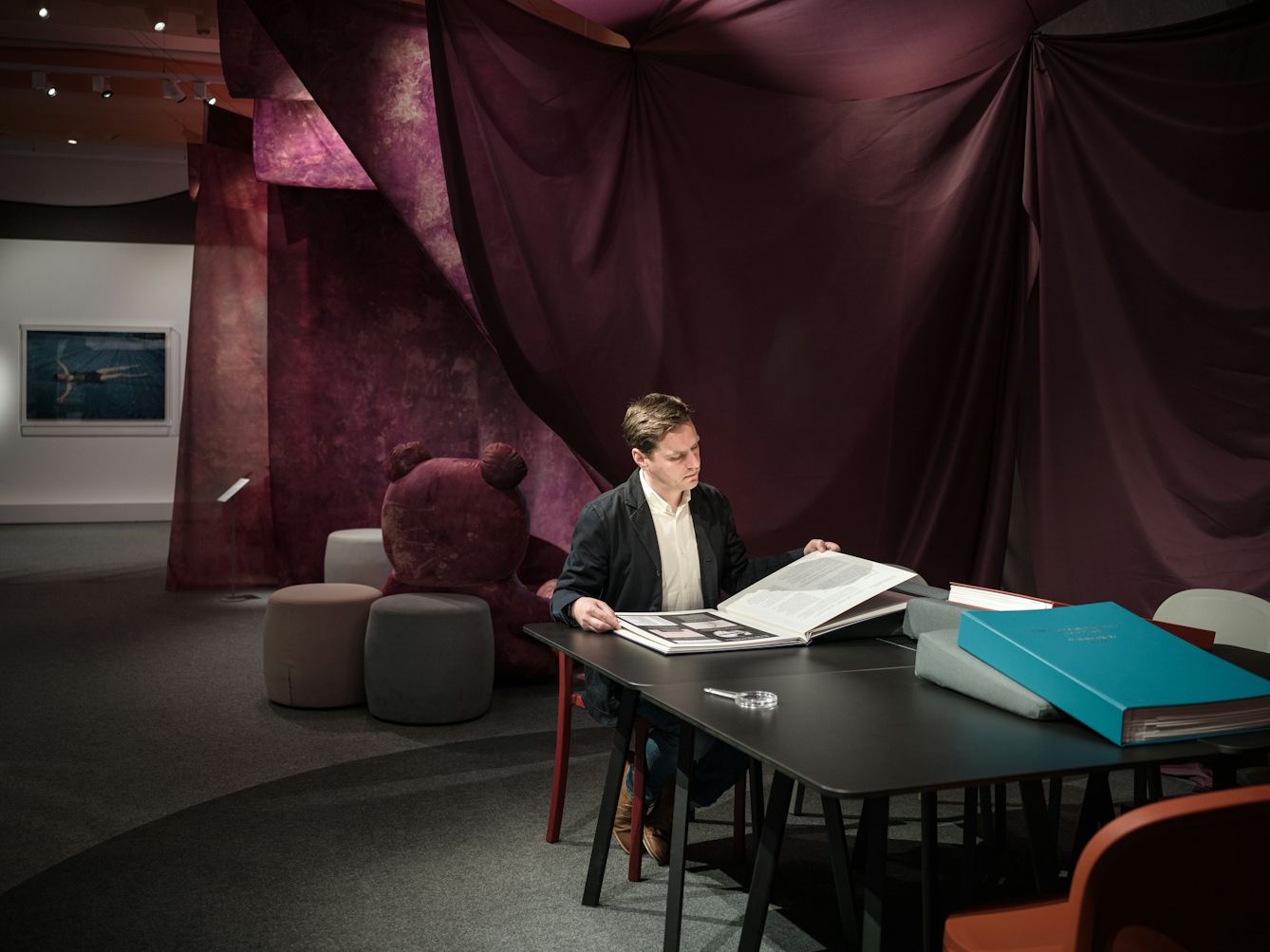 Photograph of a man sitting at a black table on a red chair leafing through a large photo book and reading the text. In the background there is the 'tent' like tie dye installation created by Oreet Ashery and the back of a life-size bear. On the table there are two other unopened books.