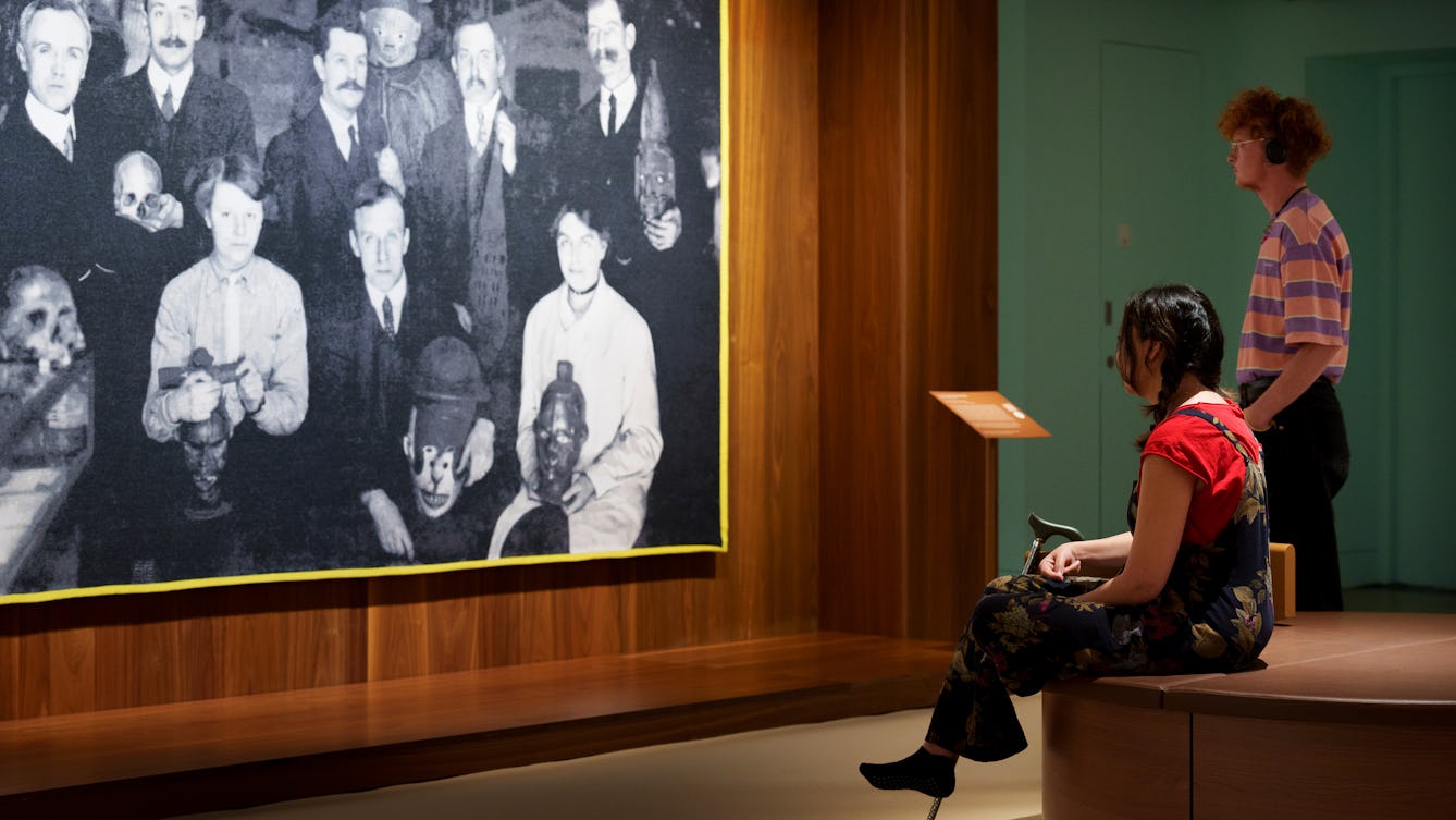 Photograph of a gallery exhibition space showing large tapestry hung on a wooden wall. The tapestry is made up of a black and white, archive photograph, from the early 20th century showing a group of white men and women gathered in a group holding museum artefacts in their hands or on their laps. They are surrounded by other museum exhibits and display cases. The tapestry is framed with a bright yellow border. Surrounding the tapestry is a wider wooden structure containing seating and alcoves. Two gallery visitors are looking at the tapestry, one standing looking at the tapestry, the other seated holding a walking stick.