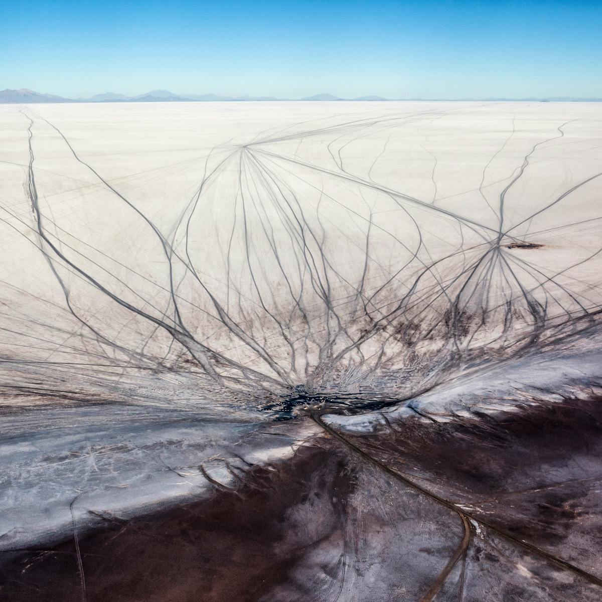 Aerial colour photograph of a large salt flat in Bolivia. The landscape is made up of a flat white expanse stretching away into the far distance where a small ridge of wavy mountains gives way to a cloudless blue sky. At the bottom of the image the white expanse meets a margin with grown earth. The surface of both the white and brown landscape are scarred with many sinewy vehicle tracks, snaking out and crisscrossing the ground all the way to the horizon.