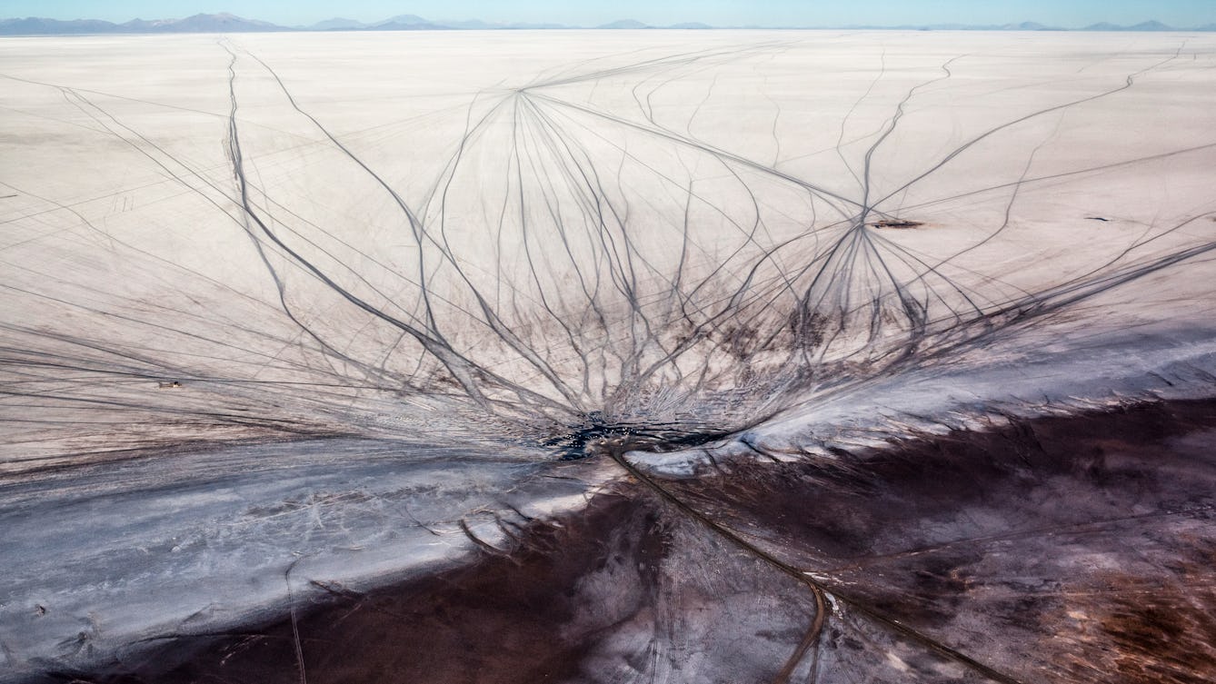 Aerial colour photograph of a large salt flat in Bolivia. The landscape is made up of a flat white expanse stretching away into the far distance where a small ridge of wavy mountains gives way to a cloudless blue sky. At the bottom of the image the white expanse meets a margin with grown earth. The surface of both the white and brown landscape are scarred with many sinewy vehicle tracks, snaking out and crisscrossing the ground all the way to the horizon.