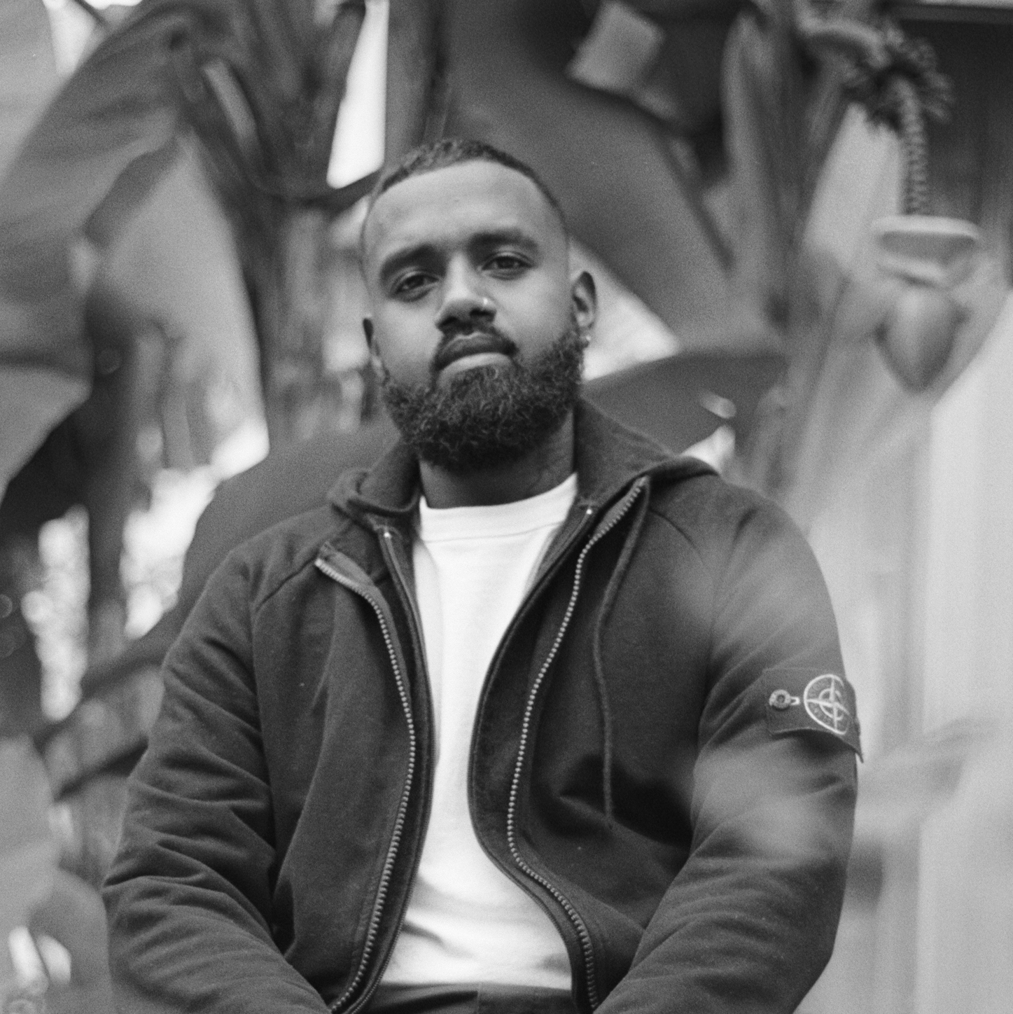 Black and white photograph of a young man with short, dark hair and a beard wearing a hoodie. The background is blurred.