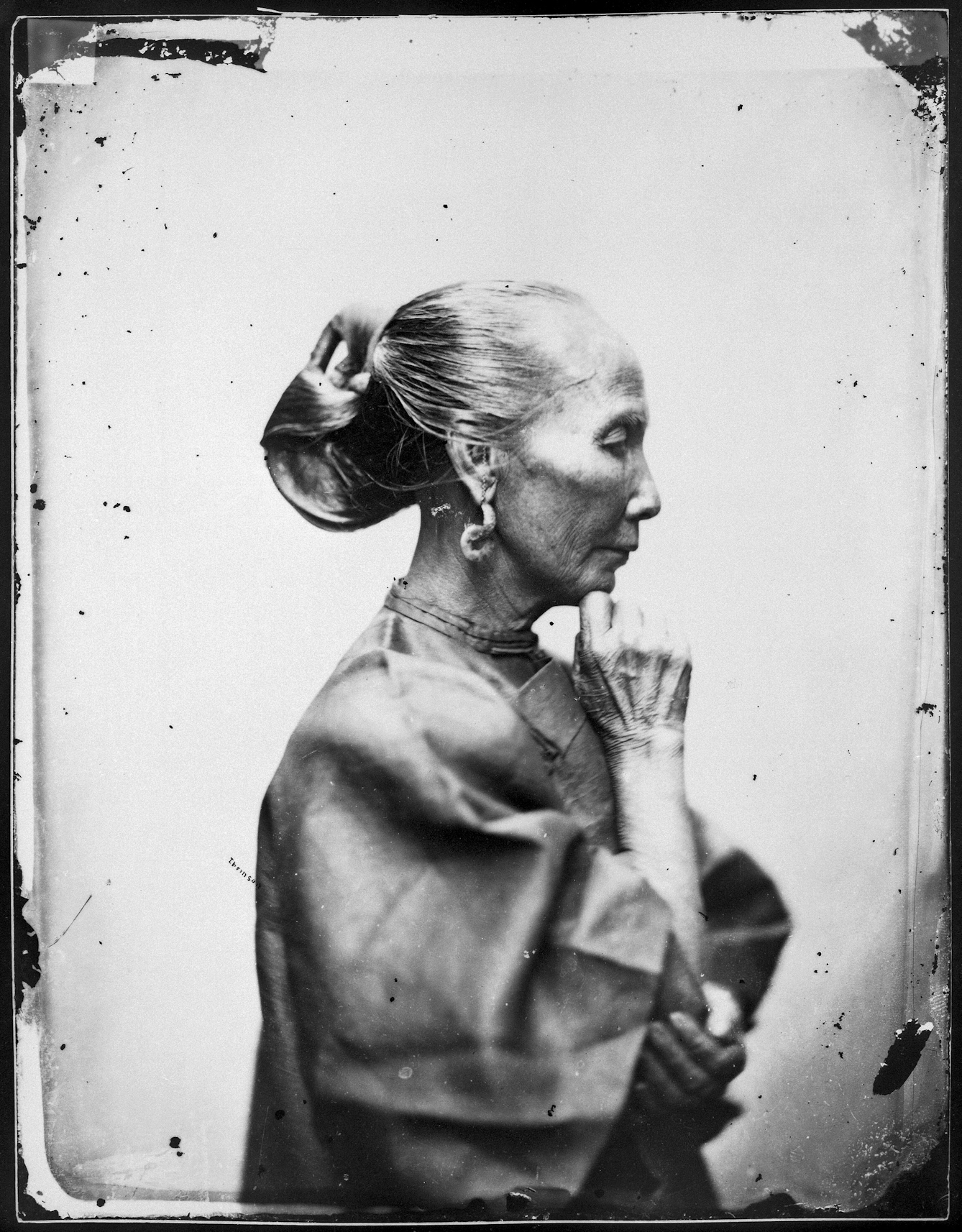 A black and white photograph of an old woman, in profile to right, resting her chin on her crooked right index finger. Her greying hair is gathered in a twist at the back. She wears large curly earrings and a garment of coarse woven material.