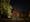 Photograph of Eltham Spiritualist Church at night. The two-storey building is set back from a main road, with a lawn and a brick wall fence. The facade has three windows, two on the ground floor and one small circular window in the apex of the roof.
