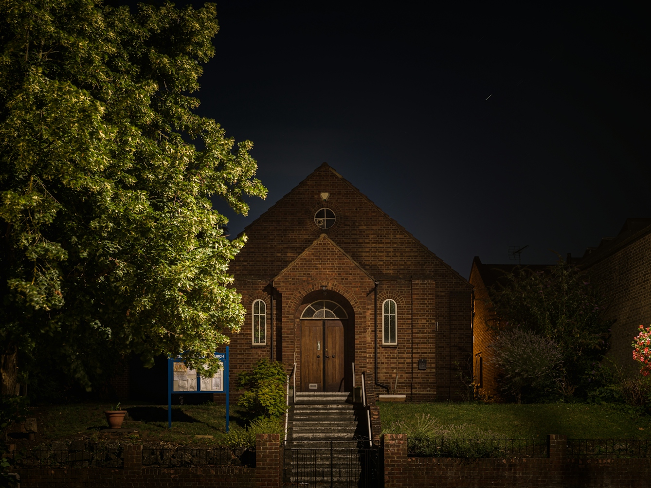 Photograph of Eltham Spiritualist Church at night. The two-storey building is set back from a main road, with a lawn and a brick wall fence.  The facade has three windows, two on the ground floor and one small circular window in the apex of the roof.