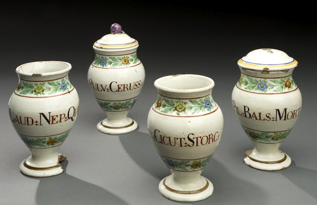 Photograph of four pharmacy vases from the early 19th Century, all cream-coloured and decorated with flowers and words indicating their contents., which include laudanum (far left) and balsamic pills (far right).