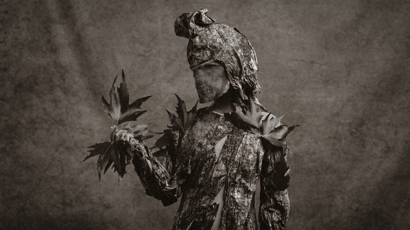 Black and white photograph of a human figure dressed in a stylised costume to imitate a tree. The costume has a bark-like surface and leaves attached to shoulders and in the hand. The figure is looking at the leaves in it's hand.