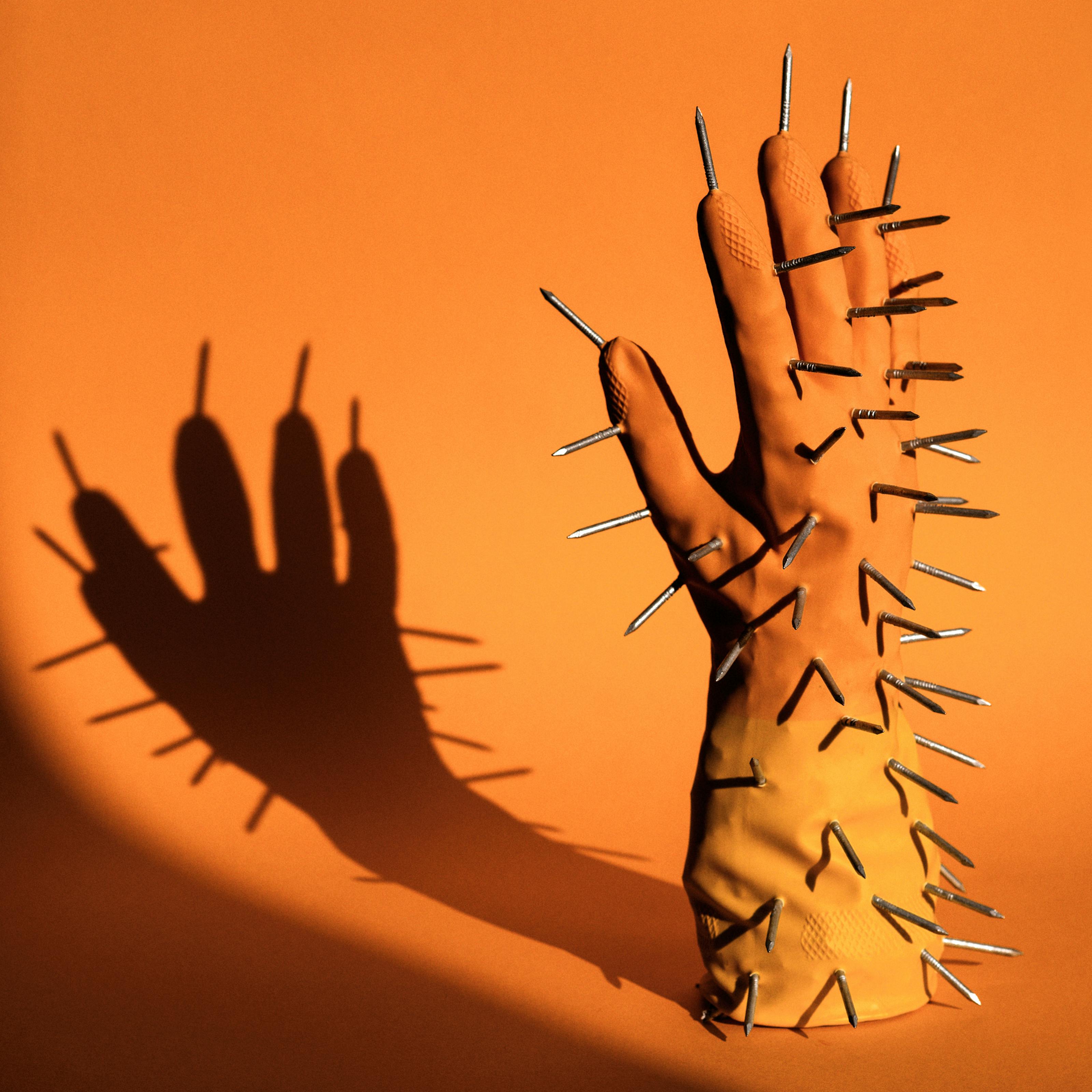 Photograph of the right hand of a pair of rubber gloves. The orange and yellow glove is standing upright, fingers into the air, on an orange background. The glove is padded so it looks as if there is a hand inside it. Sticking out of the glove all over the surface are metal carpentry nails, points outwards. The scene is lit by a hard contrasty light which is casting a long dark shadow of the glove and nails onto the background.
