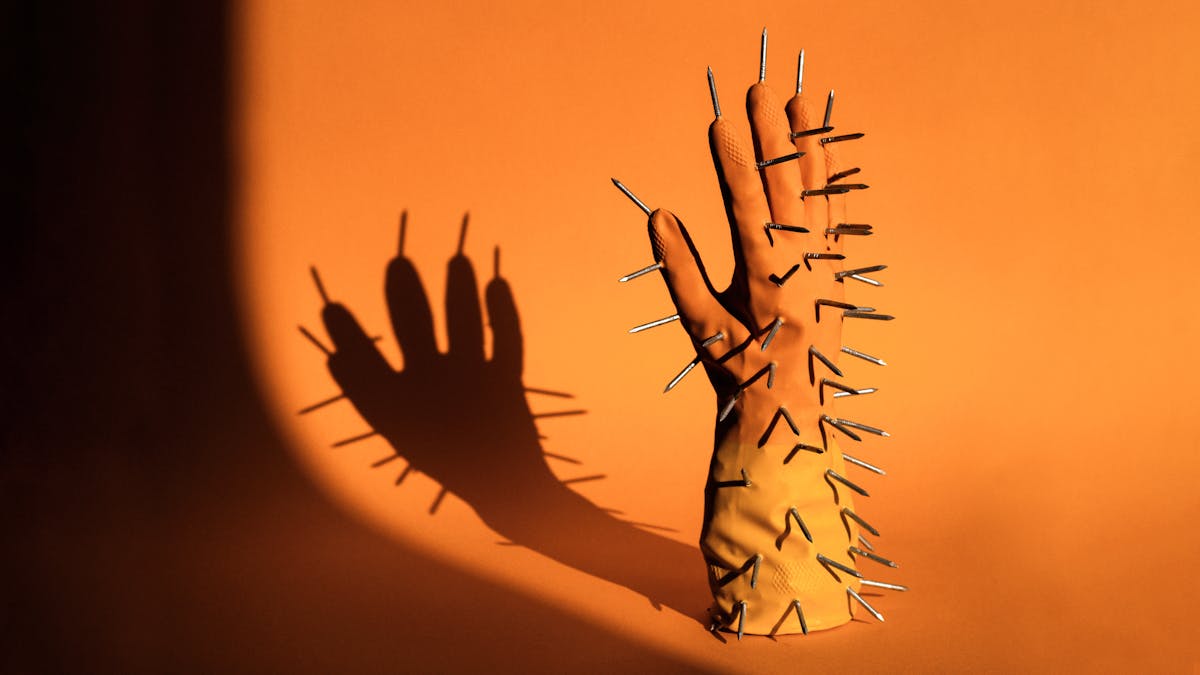 Photograph of the right hand of a pair of rubber gloves. The orange and yellow glove is standing upright, fingers into the air, on an orange background. The glove is padded so it looks as if there is a hand inside it. Sticking out of the glove all over the surface are metal carpentry nails, points outwards. The scene is lit by a hard contrasty light which is casting a long dark shadow of the glove and nails onto the background.