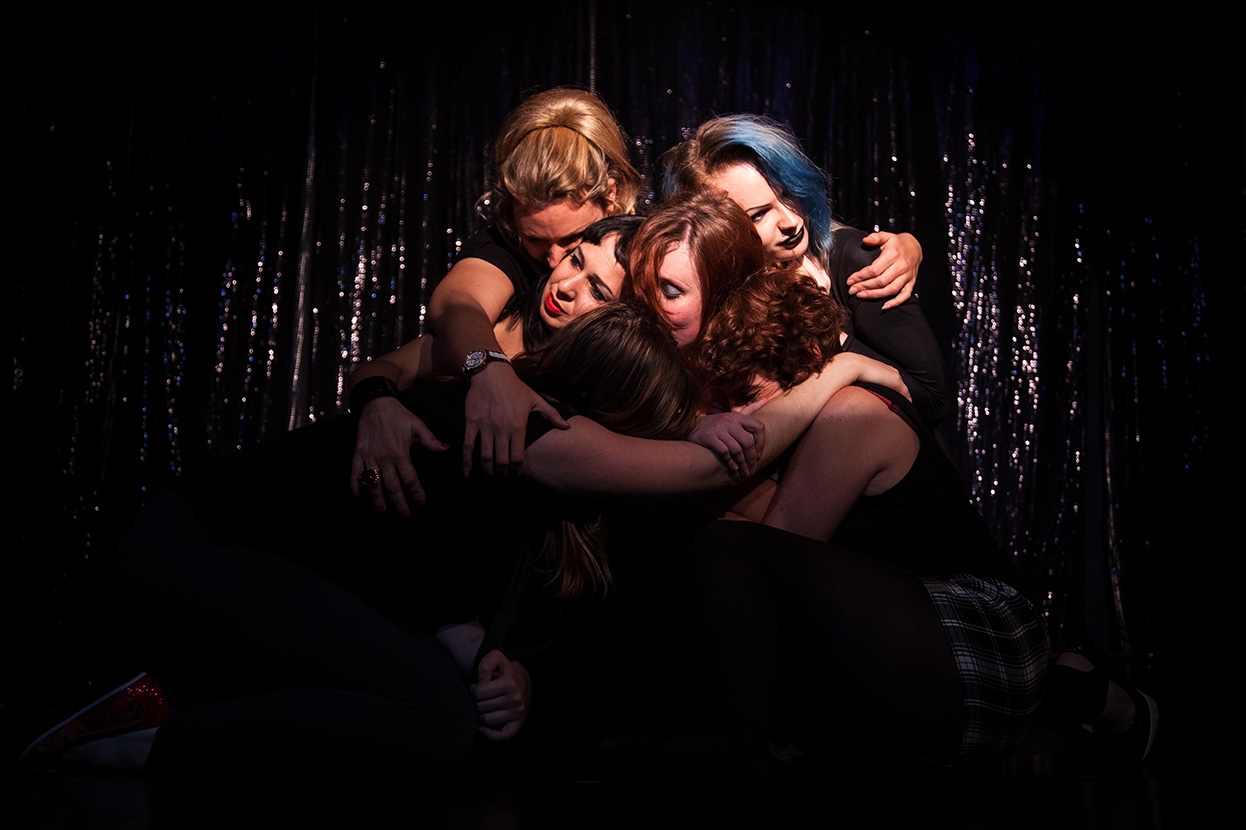 A coloured photograph group of several women with their arms around each other hugging, on a black background.