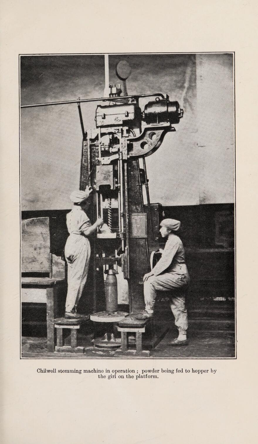 A black and white picture of two women in overalls operating a chilwell stemming machine. Accompanying text reads 'Chilwell stemming machine in operation ; powder being fed to hopper by the girl on the platform'