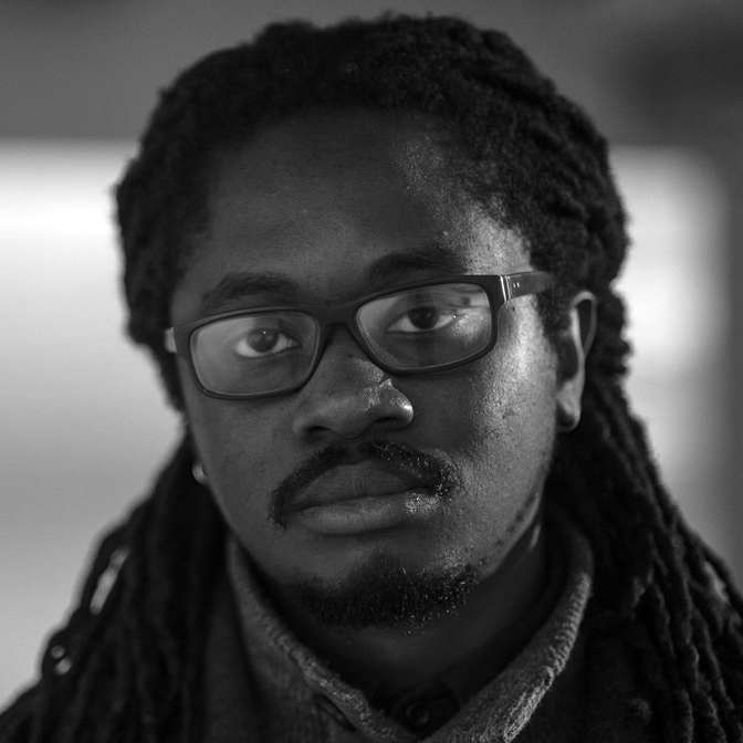 Head and shoulders photograph of a man with a tidy beard and moustache, dark-rimmed glasses and long braids.