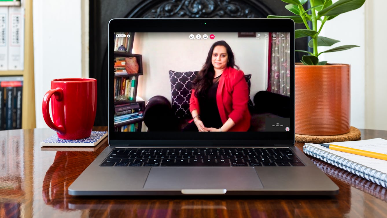 Sonia Wigh on a video call. Sonia is appearing on the laptop screen. On the desk around the laptop, there is a red mug, a small house plant and pen, and paper. 