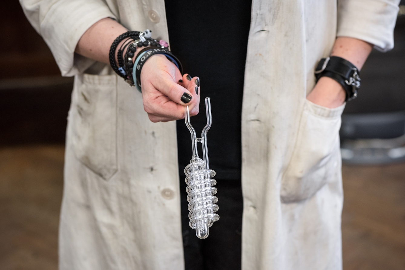 Person in a dirty white workcoat with bracelets and black nail polish holding a glass object with a spiral.