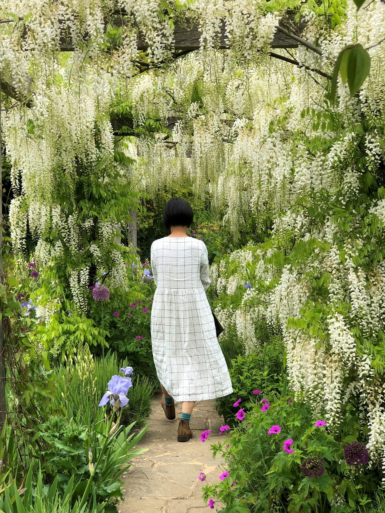 Colour photograph of a woman walking down a garden path. She is pictured from behind, wearing a long white dress and brown boots. The garden is full of flowers, which surround her on both sides and overhead.