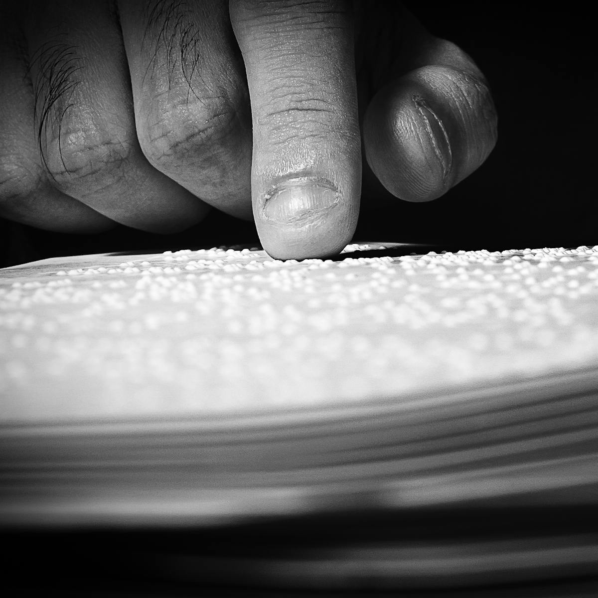 Black and white photograph of a close-up of a man's hand, whose index finger is resting lightly on white sheets of Braille. The foreground and background are dark black, casting the hand and paper sheets into a spotlight.