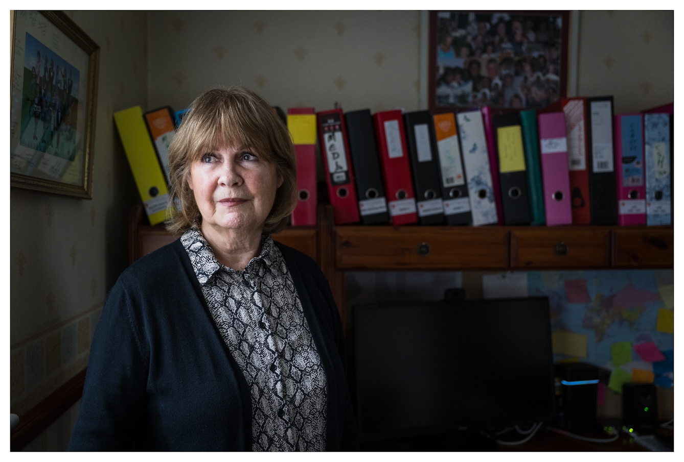 Photographic portrait of Marie Lyon stood in her study. She is looking off into distance, her face bathed in light from the window. Behind her is a long shelf full of lever arch files.