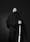 Black and white photograph of a man leaning against a wall holding the handle of a white cane in his left hand. The viewpoint is low down, looking up at the man from below. He has his right hand in his jacket pocket. He is look off into the distance, over the top of the camera. The background is a dark grey and he is stood in a pool of light, leading to him appearing as a stark figures in the frame.