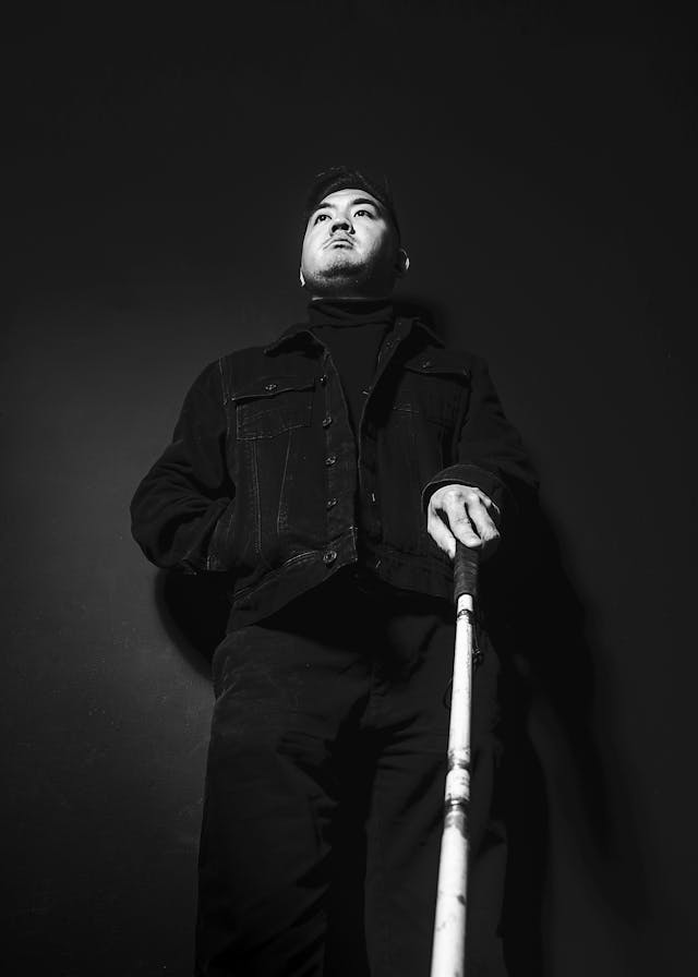 Black and white photograph of a man leaning against a wall holding the handle of a white cane in his left hand. The viewpoint is low down, looking up at the man from below. He has his right hand in his jacket pocket. He is look off into the distance, over the top of the camera. The background is a dark grey and he is stood in a pool of light, leading to him appearing as a stark figures in the frame.