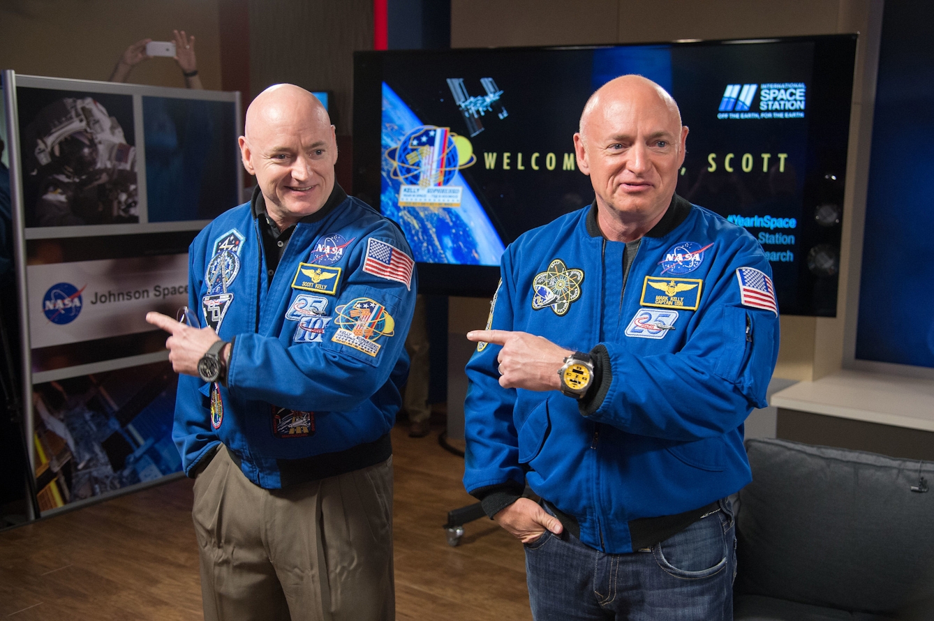 Two middle-aged bald white men who are also identical twins stand side by side in NASA jackets, each pointing to the left. Behind them is a display for NASA Johnson Space. 