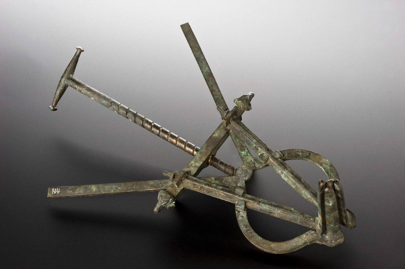 A grey metal speculum based on an ancient Roman device. Consisting of four closed metal prongs at right angles to a screw mechanism with a handle and two splayed metal arms.