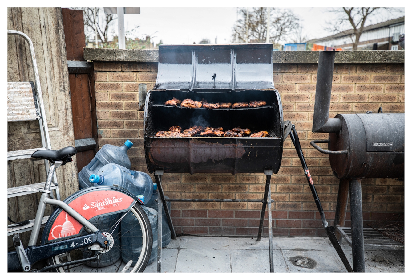 Photograph of an outdoor yard setting with paving slabs and a brick wall. In front of the brick wall are 2 large oil drum cookers, one of which has its lid open revealing the rows of chicken meat cooking inside. To the left is a bicycle and a ladder.