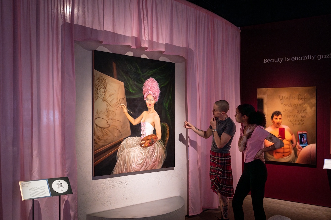Photograph of 2 gallery visitors looking at an oil painted portrait hung on a wall, surrounded by pastel pink curtains. The portrait shows a seated artist in ornate dress and pink hair, in the process of painting a similar portrait of herself within the original painting.