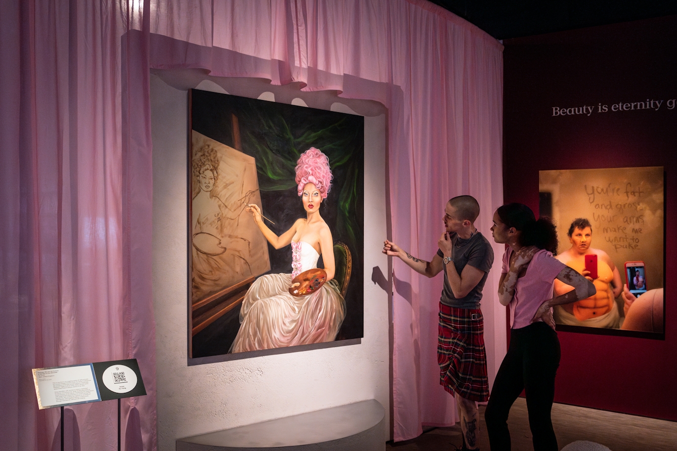 Photograph of 2 gallery visitors looking at an oil painted portrait hung on a wall, surrounded by pastel pink curtains. The portrait shows a seated artist in ornate dress and pink hair, in the process of painting a similar portrait of herself within the original painting.