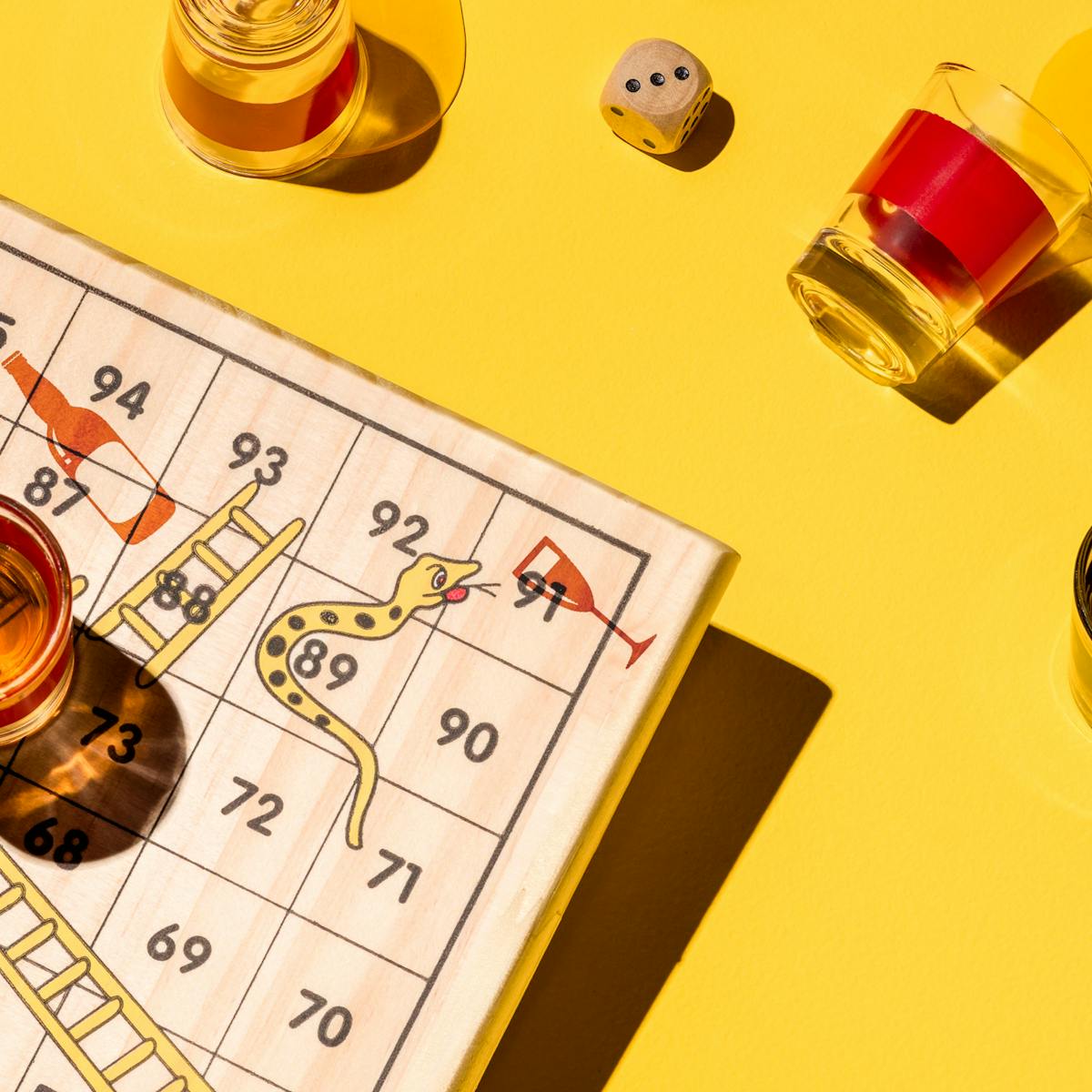 Photograph of a board game of snakes and ladders made of wood, sitting on a yellow background. The snakes on the board game are drinking alcohol. Surrounding the board are five shot glasses, three of which are brimming with alcohol, and two which have been spilled. In the centre right of the frame there are two dice reading the numbers five and three.