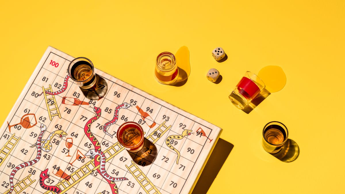 Photograph of a board game of snakes and ladders made of wood, sitting on a yellow background. The snakes on the board game are drinking alcohol.  Surrounding the board are five shot glasses, three of which are brimming with alcohol, and two which have been spilled.  In the  centre right of the frame there are two dice reading the numbers five and three.
