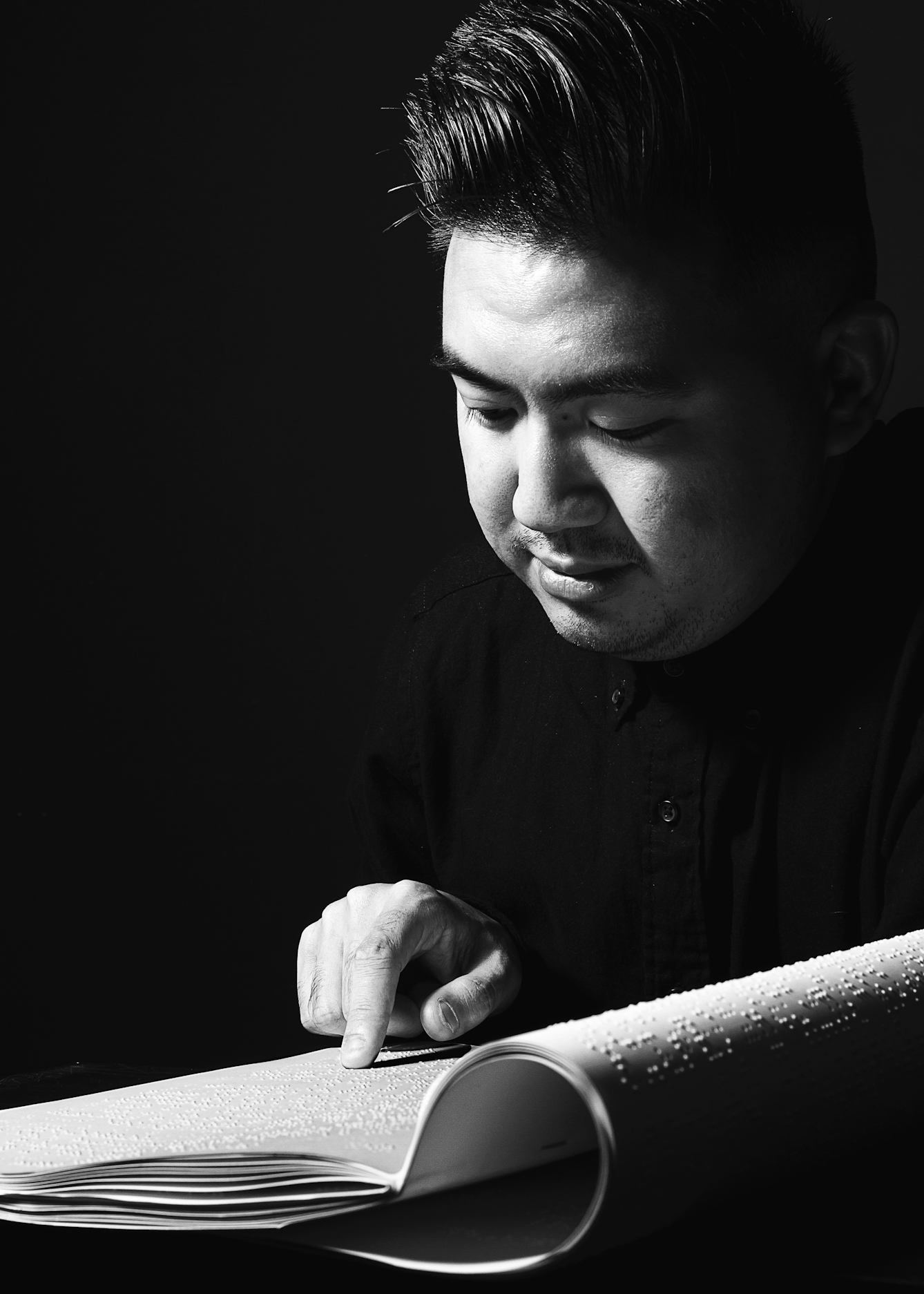 Black and white photograph of a man seated at a table with the index finger of his right hand resting lightly on white Braille sheets. His face is angled down towards the pages. The man's face, hand and the papers are spotlit from above, casting the foreground and background into darkness.