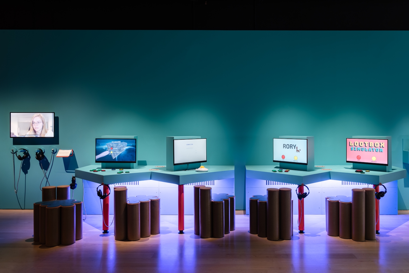 Photograph of an exhibition space showing 4 display screens mounted on tabletops with 5 chairs made out of brown cylinders. Hanging from the tables are sets of headphones. On one screen is the word, Rory, on another are the words, Lootbox simulator.