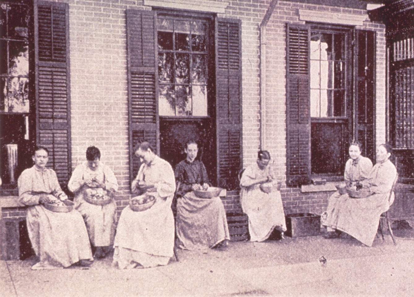 A group of women seated outside a large building, peeling vegetables into bowls on their laps
