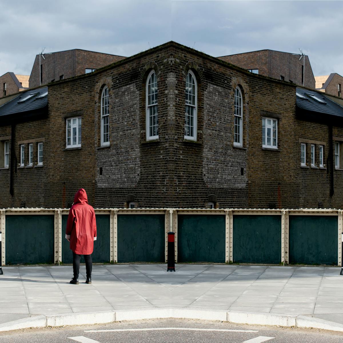 Photographic panorama showing a street scene with part of a road, a pavement, the handrail of a bridge and in the distance industrial brick buildings. The panorama is mirrored down its vertical centre line except that a small human figure in a red coat standing on the pavement looking away from the camera appears in the left half, but not in the right half.