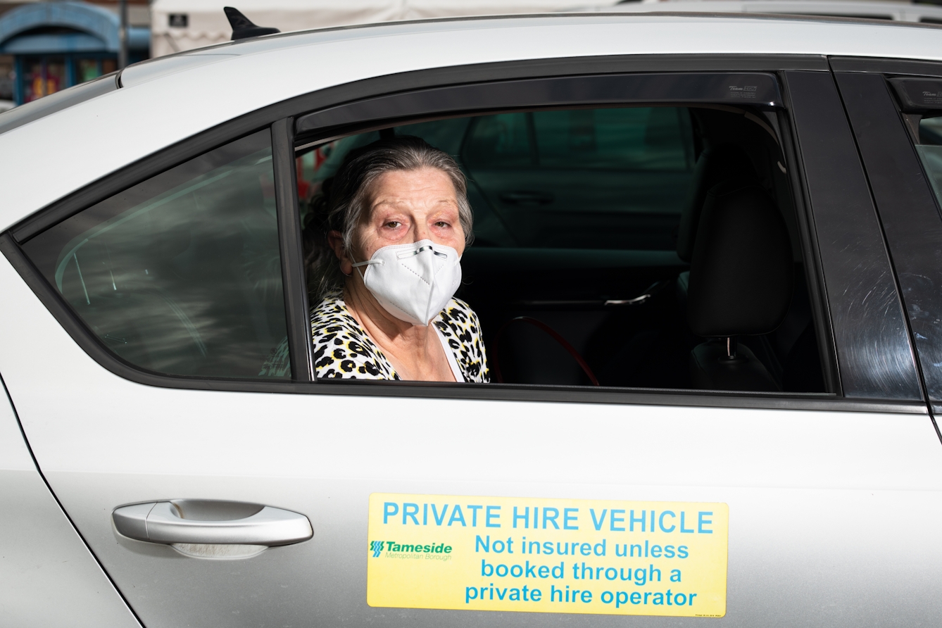 Photographic flash lit portrait through the open side window of a stationary car. The car is silver and the front of the car is facing to the right. On the door is a large sign with the words, 'Private Hire Vehicle Not insured unless booked through a private operator'. Sat in the rear passenger seat is a woman, looking to the camera. She is wearing a white face covering. In the background is the out of focus scene of a building and other cars.