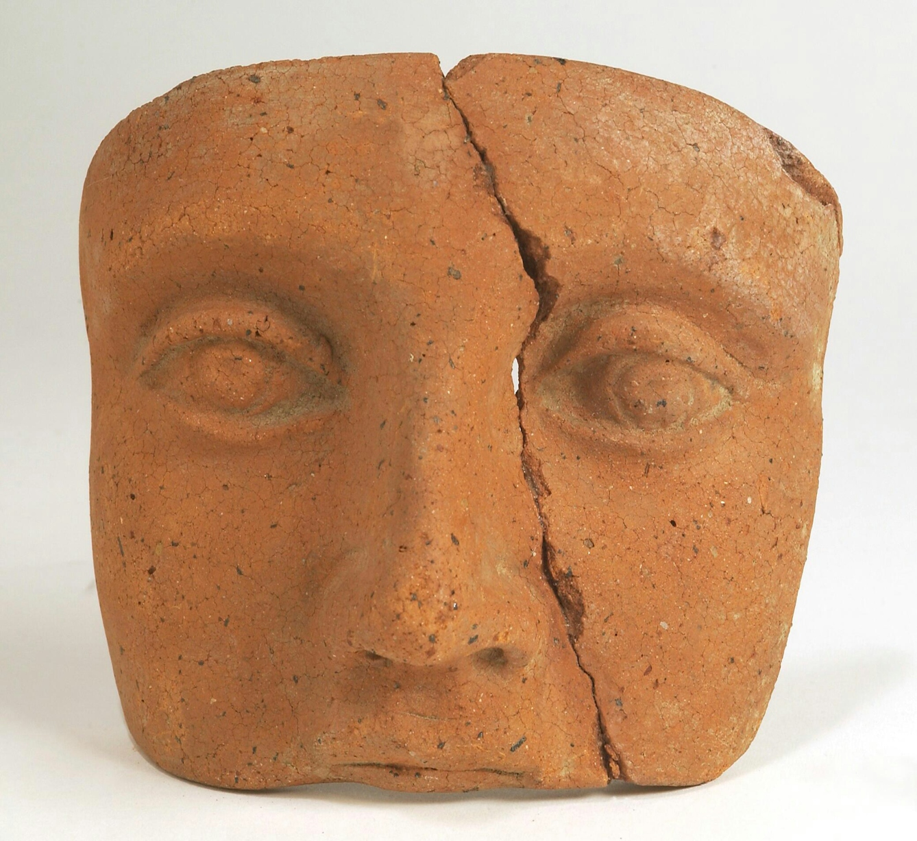 Photograph of a clay Roman votive offering. The votive is in the shape of a human head, from the upper lip to the forehead. The votive has cracked vertically at a slight diagonal, meaning the votive is in two separate parts. 