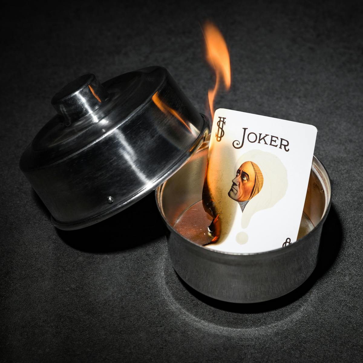 Photograph of a burning joker card in a silver dish. On the card is the face of the 1920s mind reader, Alexander.