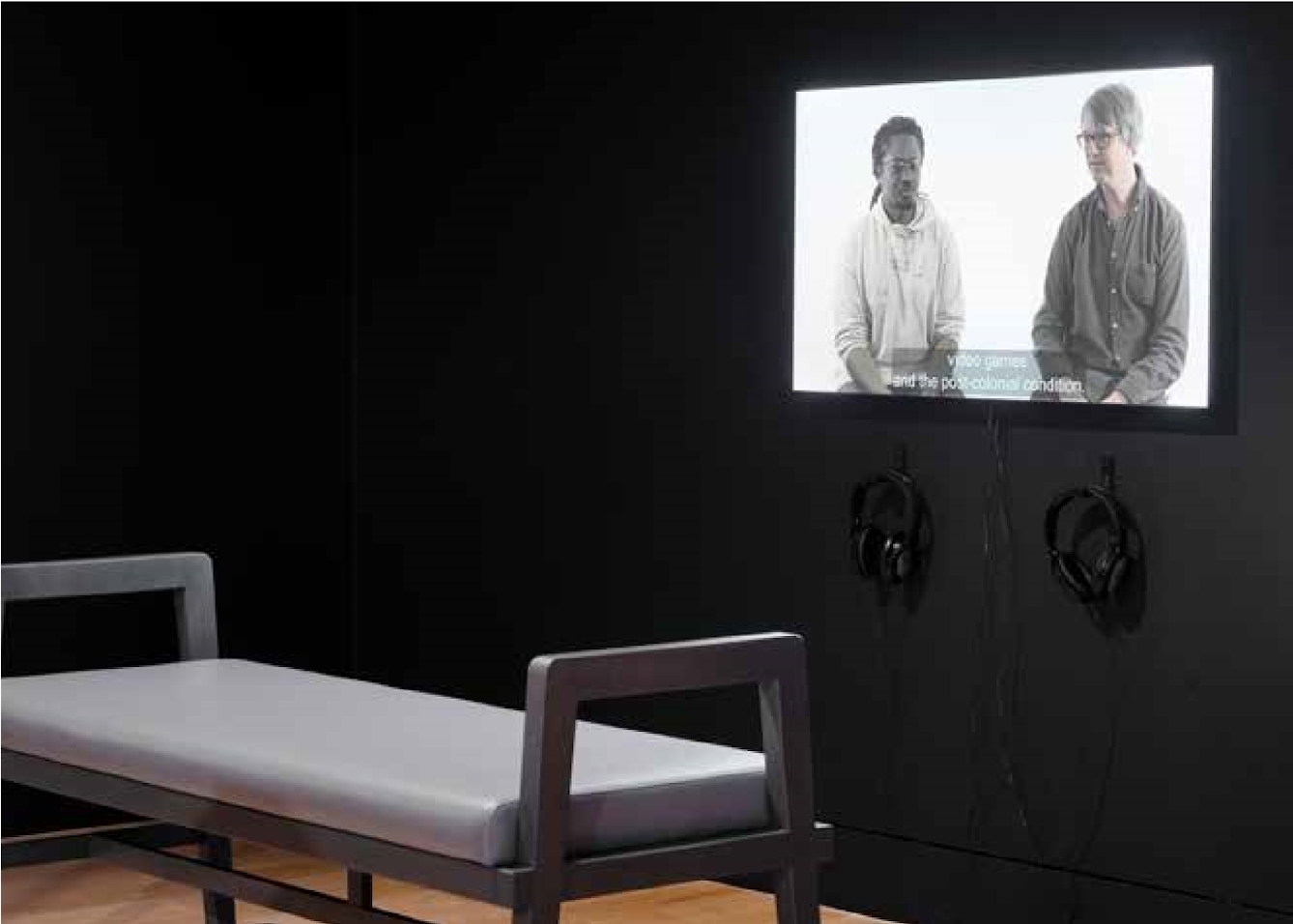 A video screen with two men sat side by side hangs on a black painted wall with two headphones hanging on the wall beneath the screen. A long bench is placed immediately infront of the screen.